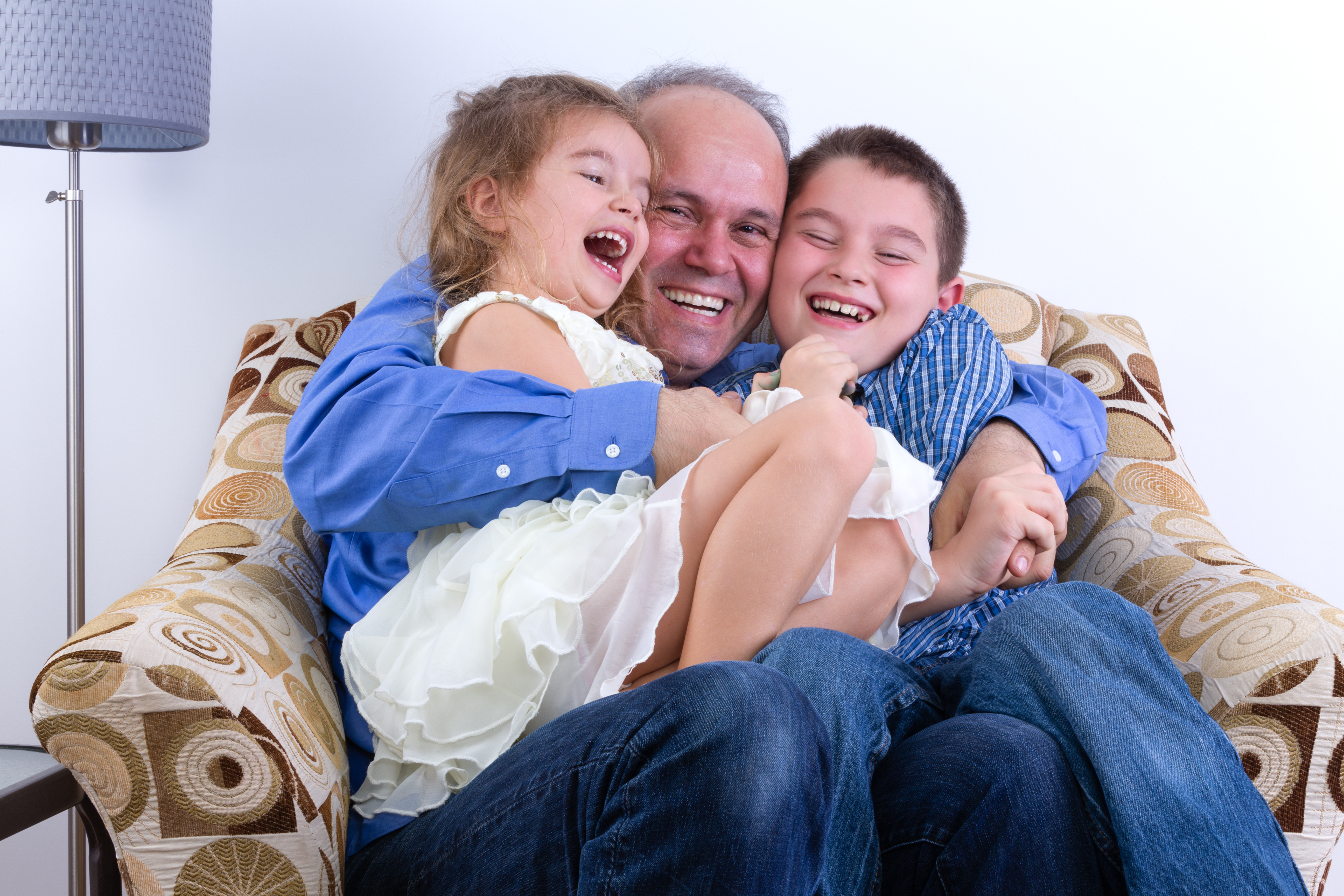 Middle-aged single father with two laughing young kids, a girl and boy, on his lap sitting in an armchair enjoying a moment of fun and hilarity as a family
