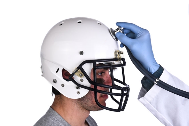 mild traumatic brain injuries in sports - A football player wearing a helmet with doctors hand holding a stethoscope on the crown of the helmet