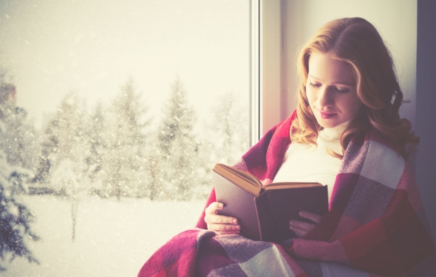 reducing winter bills and expenses - happy girl reading a book by the window in the winter