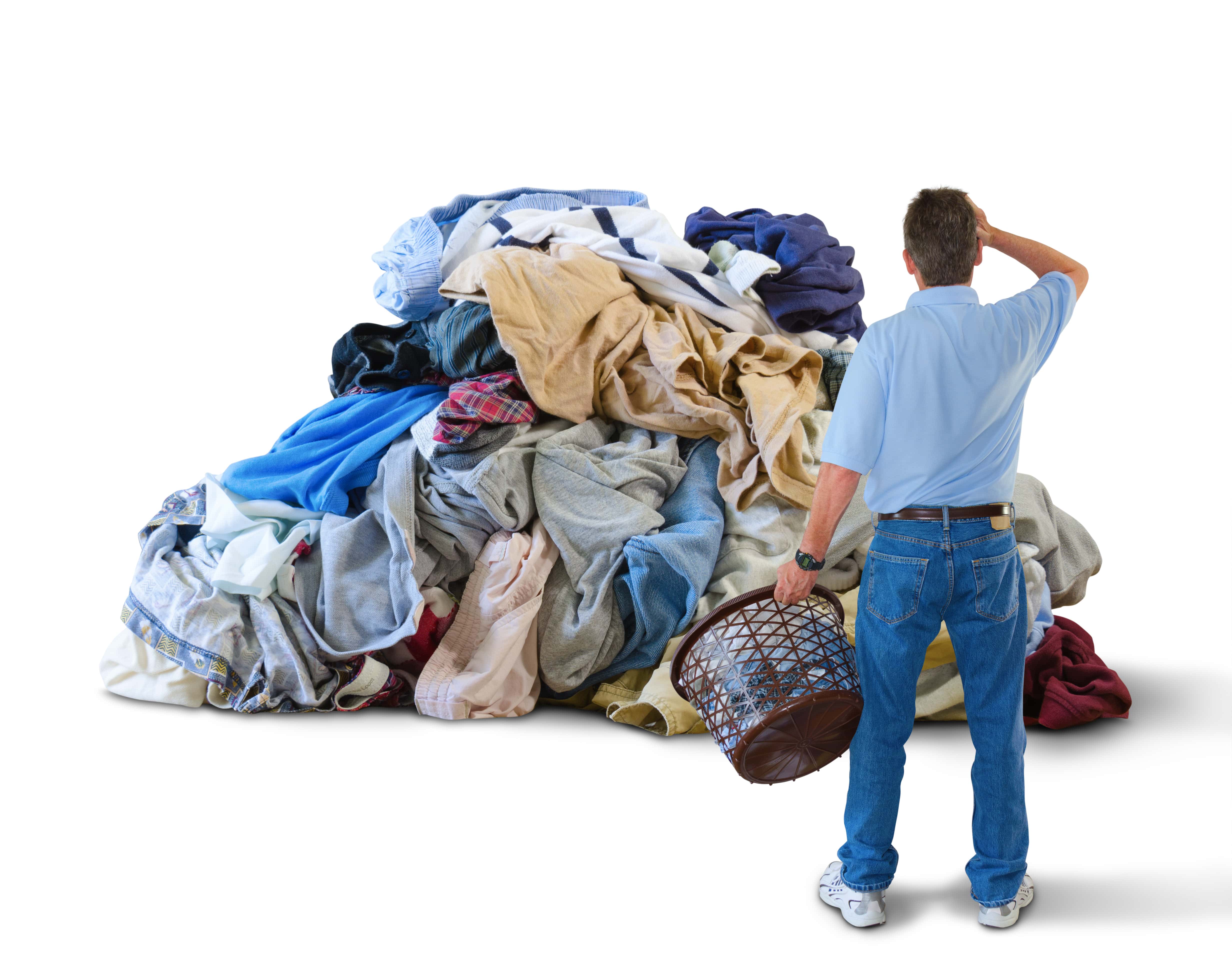 A distraught man with his hand to his head and a laundry basket in his hand is standing in front of a giant pile of dirty close.