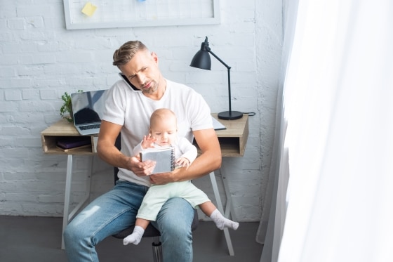 overcoming boredom before it overcomes you -busy father sitting on chair with notebook, talking on smartphone and holding baby daughter at home