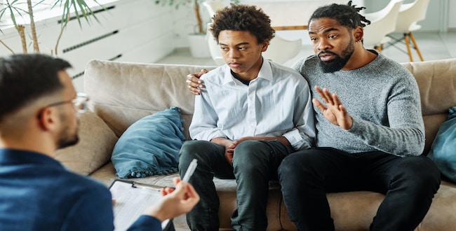 Stepfather with his teenage stepson at a meeting with a psychologist discussing mental health. family sitting on the sofa in the therapist's office.