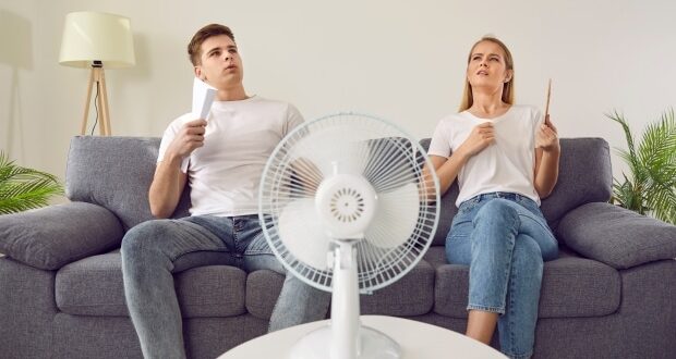 Couple at home suffering from hot sultry sweltering summertime weather. Sweaty exhausted people waving sheets of paper sitting on sofa with electric fan in overheated house.