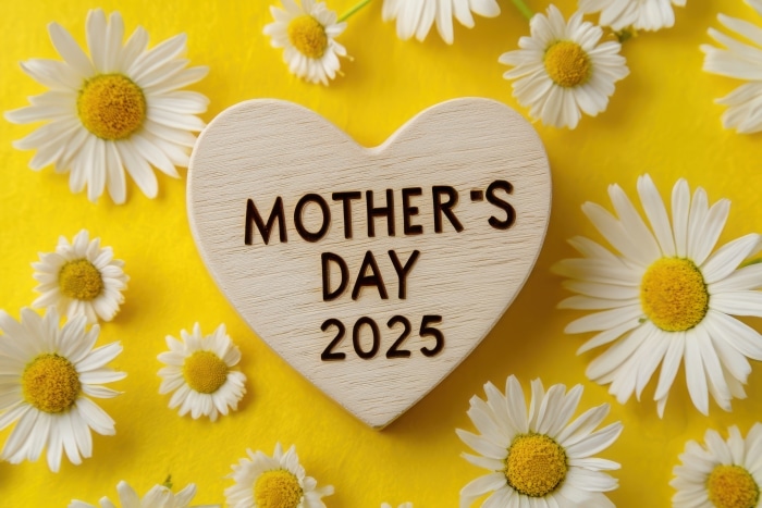 wooden heart with text "MOTHER'S DAY" and the year '2025' on a yellow background, surrounded by daisy flowers.