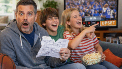 March Madness with your stepkids - Stepdad sitting confidently with a bracket while stepkids laugh and point at a TV showing an upset game.